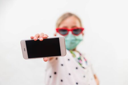 Little blond girl wearing disposable mask and sunglasses holding mobile phone over white backgroundの写真素材