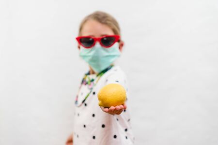 Little blond girl wearing disposable mask and sunglasses holding yellow lemon vitamin C source over white backgroundの写真素材