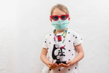 Little blond girl wearing disposable mask and sunglasses and holding microscope device over white backgroundの写真素材