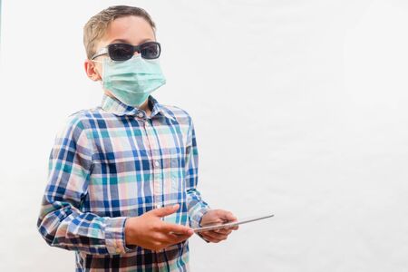 School boy wearing disposable mask and sunglasses holding tablet over white background. Homeschooling conceptの写真素材