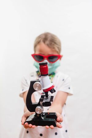 Little blond girl wearing disposable mask and sunglasses and holding microscope device over white backgroundの写真素材