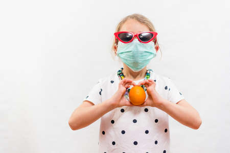 Little blond girl wearing disposable mask and sunglasses holding an orange vitamin C source over...