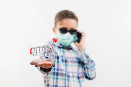 Little school boy wearing disposable mask and sunglasses and holding small shopping cart and mobile phone over white backgroundの写真素材