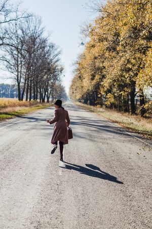 Young redhead lady woman in pink vintage coat and hat with suitcase in retro style runningaway along a park road with golden yellow autumnal trees. Outdoor autumn garden relaxation, travel conceptの写真素材