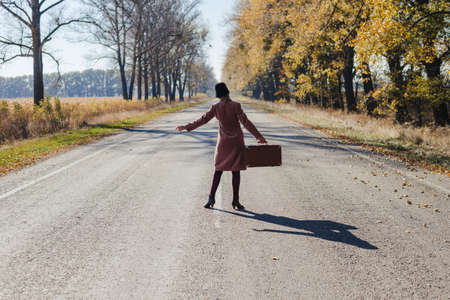 Young redhead lady woman in pink vintage coat and hat with suitcase in retro style walking away along a park road with golden yellow autumnal trees. Outdoor autumn garden relaxation, travel conceptの写真素材