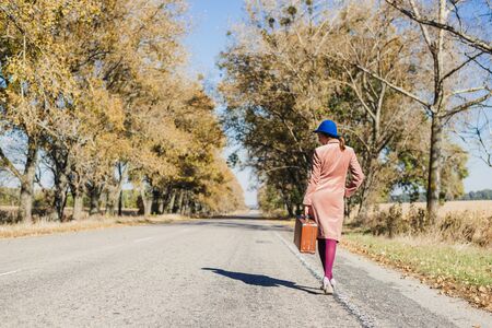 Young redhead lady woman in pink vintage coat and hat with suitcase in retro style walking away along a park road with golden yellow autumnal trees. Outdoor autumn garden relaxation, travel conceptの写真素材