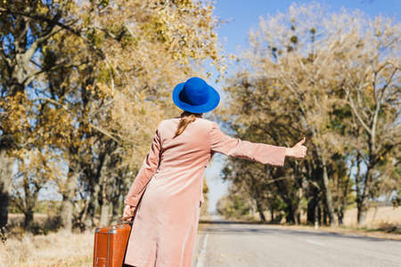 Young redhead lady woman in pink vintage coat and hat with suitcase in retro style walking away along a park road with golden yellow autumnal trees. Outdoor autumn garden relaxation, travel conceptの写真素材