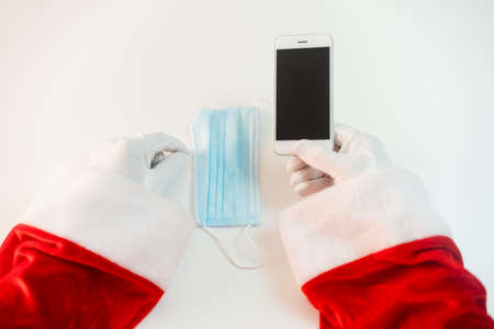 Santa Claus in a medical mask with a smartphone. Online greetings, ordering services for Christmas and new year. Social distance in the coronavirus epidemic, a new reality. Copy spaceの写真素材