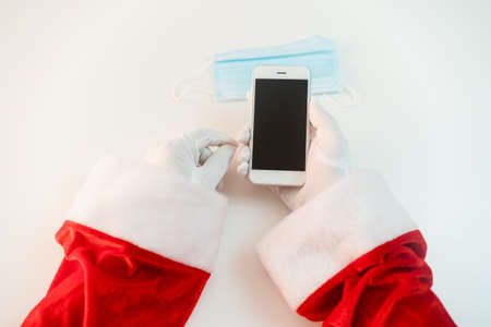 Santa Claus in a medical mask with a smartphone. Online greetings, ordering services for Christmas and new year. Social distance in the coronavirus epidemic, a new reality. Copy spaceの写真素材