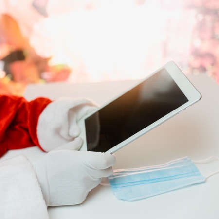 Santa Claus in a medical mask with a tablet. Online greetings, ordering services for Christmas and new year. Social distance in the coronavirus epidemic, a new reality.の写真素材