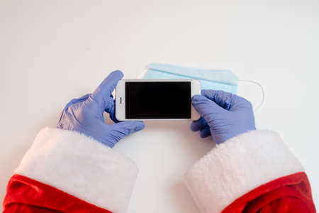 Santa Claus hands holding smart phone and a medical mask over white table background.