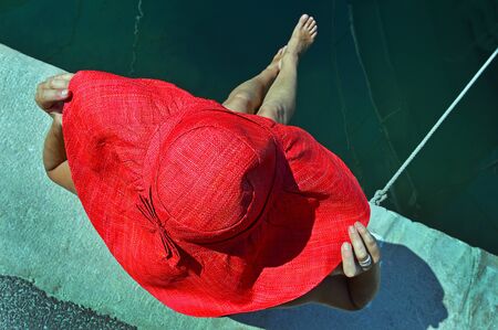 A young woman in a red beach hat sits on the pier near the yachtの写真素材