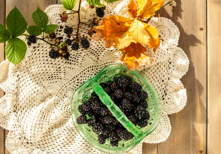 Vintage green glass basket dish full of juicy blackberries with oak leaves and blackberry sprigs on crocheted lace doily cloth on wooden table.の写真素材