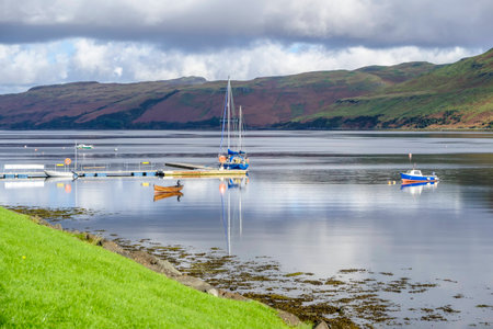 Boats and their reflections on the beautiful calm waters of Loch Harport at Carbost Isle of Skye Scotland with hills of purple heather beyond under a cloudy skyの写真素材