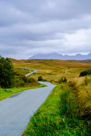 A narrow single track road winds throuth the beautiful barren landscape of Isle of Skye Scotland with the peaks of Cuillin mountain range reaching into the clouds on the horizonの写真素材