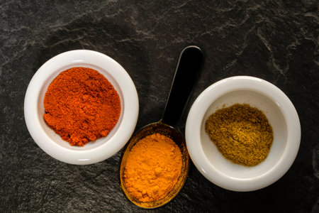 Vibrant colors of spices in small white dishes and black measuring spoon lay flat on textured black slate. Turmeric, cumin and paprika.の写真素材