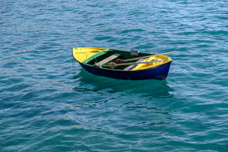 Small colorful row boat and oars moored in the clear blue water of the Atlantic Ocean with reflection on the waves and ripples.の写真素材