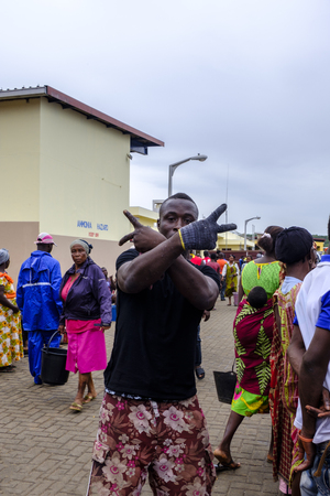 SEKONDI TAKORADI, GHANA â 10 APRIL 2018: Young male worker wearing one glove at busy Bosomtwi Sam Fishing Harbour fish market crosses his arms and makes hand gesturesのeditorial素材
