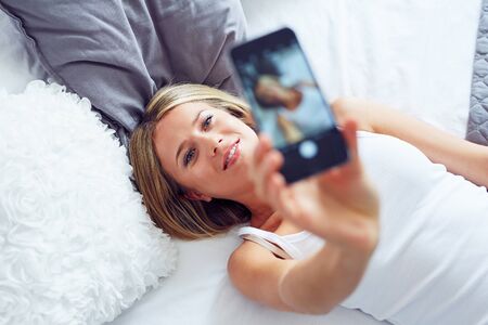 Beautiful young woman lying on the bed and making selfie photo on smartphone