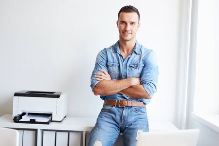 Young businessman standing by his desk in office with crossed armsの写真素材