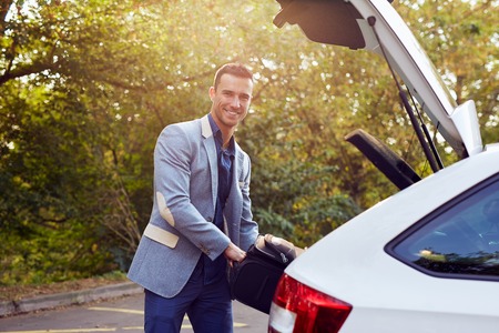 Happy man stands by the car and puts the suitcase in the trunk