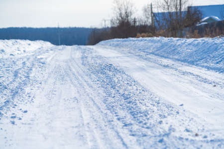 Rural road in winter covered with rolled snow on the background of snow-covered fields and forests with a clear blue skyの写真素材