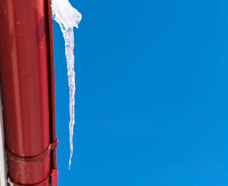 A large transparent icicle against a clear blue sky hangs from a red drainpipe in winter on the eve of springの写真素材
