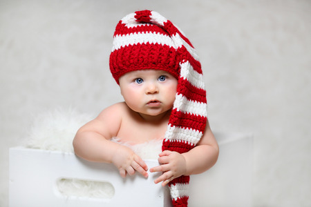 a christmas baby sitting in a white wooden box wearing a red and white striped long-tailed gnome hatの写真素材