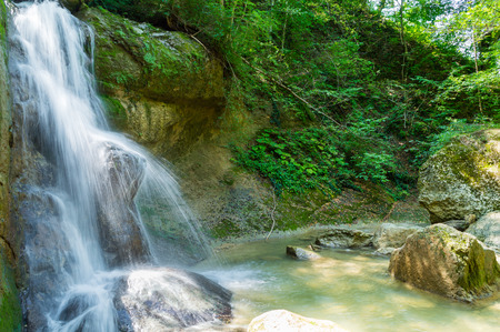 Waterfall from a cliff in the mountains. Cascade on the river. Stones covered with moss in the lake with clear water. Water in Motion. Republic of Adygea, Southern Russia.の写真素材