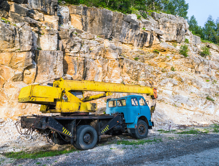 Old yellow automobile crane with blue cab and telescopic boom in transport position near with rocky mountain, left rear coner viewの写真素材