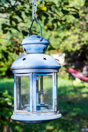 Light blue garden candle lamp hanging on a tree. In the background, the garden in the morning sun. Background blurred.の写真素材