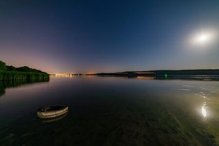 Lake or river sand shore with trees and dark blue starry sky and city light on background. Tranquil nature night landscape.の写真素材
