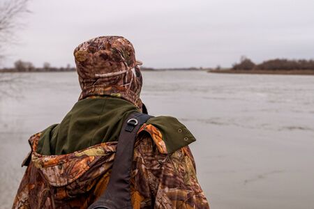 Man in camouflage trousers and jacket with hood catches fish on river in a cloudy day.の写真素材