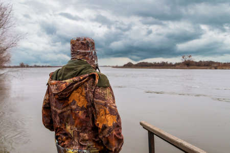 Man in camouflage trousers and jacket with hood catches fish on river in a cloudy day.の写真素材