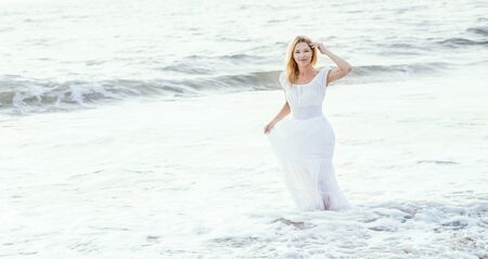 Attractive happy mature tourist blonde woman in long white dress on asian sand tropical beach smiling joyful near water. Romantic travel vacation, active lifestyle, independent and confident person with copyspace banner.の写真素材
