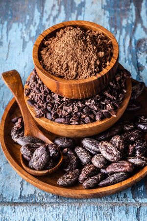 Organic raw cocoa beans, nibs and powder in wooden bowls on blue rustic background close-up vertical shot selective focus.の写真素材