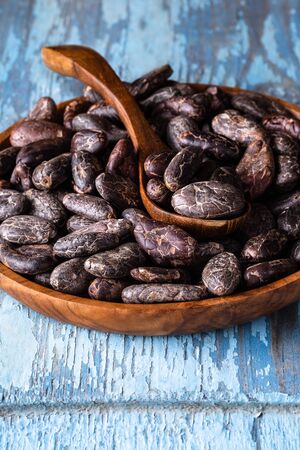Cocoa beans in wooden bowl on blue rustic background close-up vertical shot selective focus.