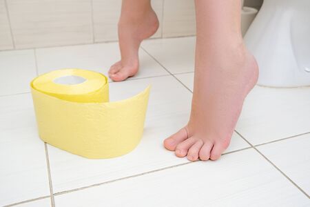 Cropped legs of a boy sitting on a toilet with a roll of yellow toilet paper on a floor, digestive...