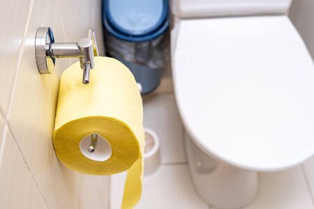 A roll of yellow toilet paper hanging on a wall fixture in a rest room with a toilet bowl on background, digestive problems concept.の写真素材
