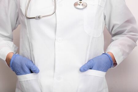An unrecognizable doctor in white uniform and blue gloves with a stethoscope on neck holding hands...