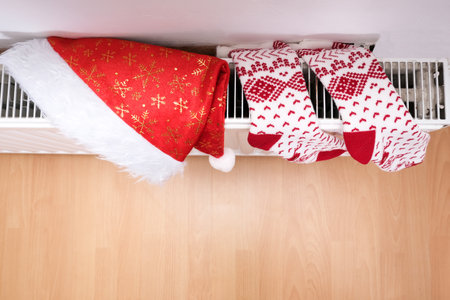 White and red christmas socks and santa hat hanging on warm central heat radiator, winter holiday home mood.の写真素材