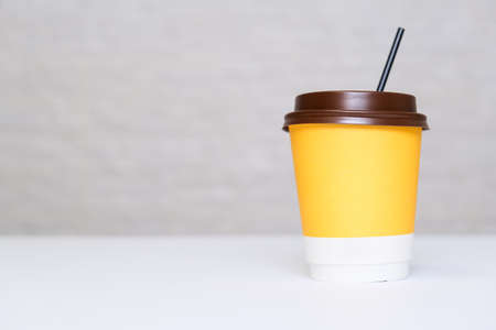 A yellow paper disposable takeout coffee cup with a brown cap and a straw on white background against light brick wall with copy spaceの写真素材