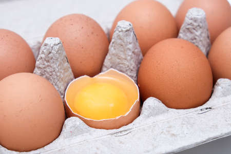 A brown chicken egg is half broken among other eggs in a carton trayの写真素材