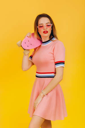 Portrait of happy smiling blonde girl hold pink skateboard on yellow background. People summer vacation rest lifestyle concept. Posing in pink short dress and sunglassesの写真素材