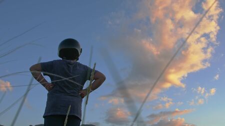 boy in a motorcycle helmet with his hands resting on his hips admiring the sunset in the mountains view from the backの写真素材
