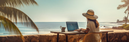 Rear view of young woman, female freelancer in straw hat working on laptop, while sitting on the tropical sandy beach.の素材