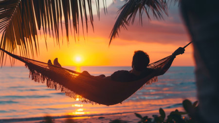 A man enjoys calm, lies in a hammock on the background of the ocean and sunset. Handsome guy meets the sunset on the beach of a tropical.の素材