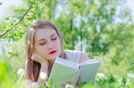 Young pretty woman in white top with long hair lies on the grass surrounded by flowers in apple tree garden and reading book on sunny day. Copy space for your textの写真素材