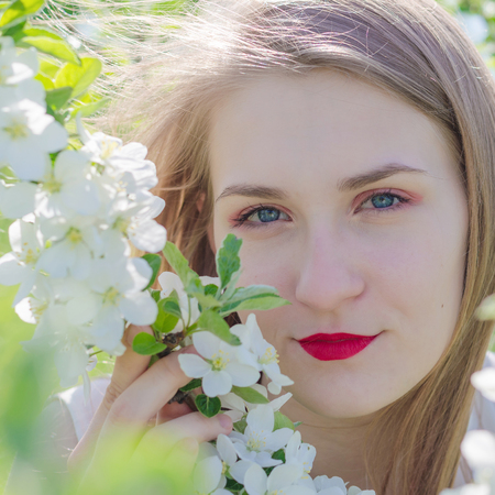 Portrait of a young beautiful woman in white top standing among blooming apple trees on a sunny day. Close-up with space for text.の写真素材