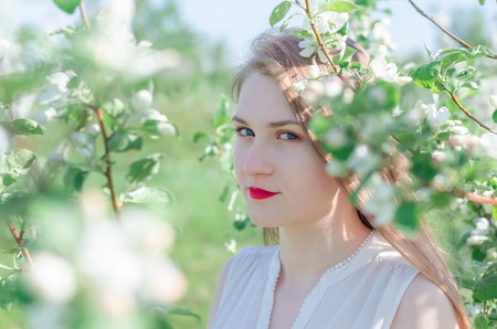 Portrait of a young beautiful woman in white top standing among blooming apple trees on a sunny day....